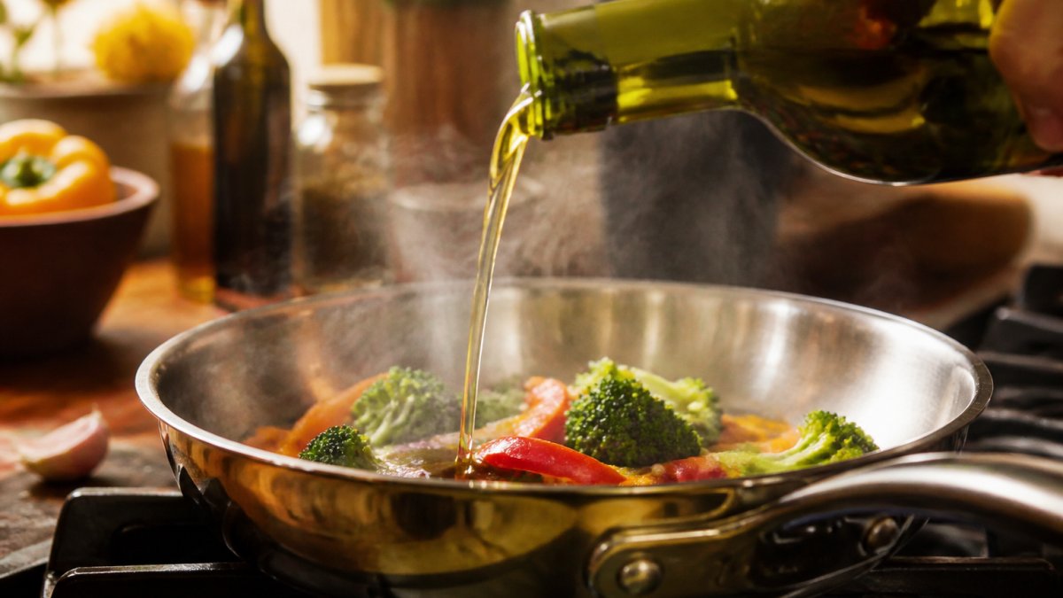 Close-up of organic oil being poured into a stainless steel pan with steam rising as vegetables prepare to sear, warm kitchen lighting with golden tones
