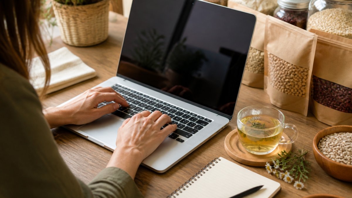 Person's hands typing on laptop keyboard while searching the USDA Organic Integrity System, with a cup of herbal tea and notepad on wooden desk beside the computer